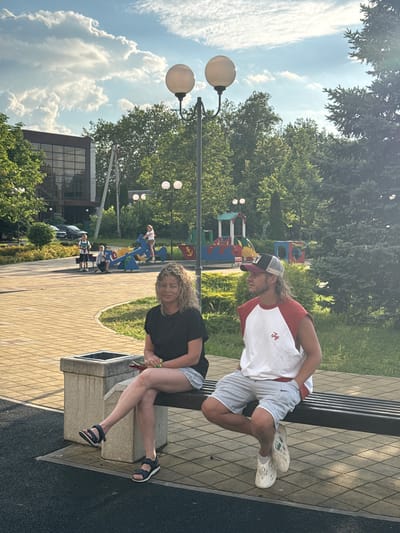 In Kurganinsk, Russia, a woman with curly blonde hair sits on a bench in what appears to be a park or playground area, facing left. She is wearing a black t-shirt and light-colored shorts, with dark sandals on her feet. To her right, a man with long blonde hair and a baseball cap turned backward is also seated on the bench, facing left. He is wearing a white tank top with red sleeves and gray shorts, with light-colored, perforated shoes. In the background, a playground with colorful equipment, including a seesaw, is visible, along with several children playing. Trees and a building with large windows are also present in the environment, under a partly cloudy sky.
