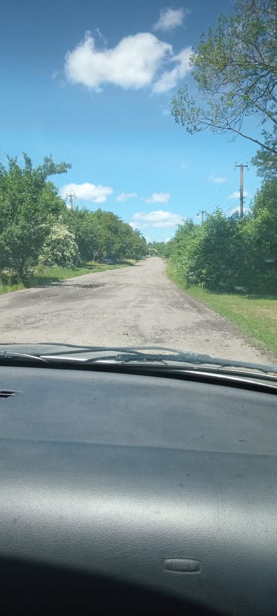 The image, taken from inside a vehicle, shows a damaged asphalt road leading into the distance. The road is lined with dense green trees on both sides. Utility poles with overhead wires are visible along the right side of the road. The sky is blue with scattered white clouds. A portion of the car's dashboard, including the airbag cover, is visible in the foreground. The scene appears to be a rural or semi-rural area. The location is specified as Koviahy, Ukraine.