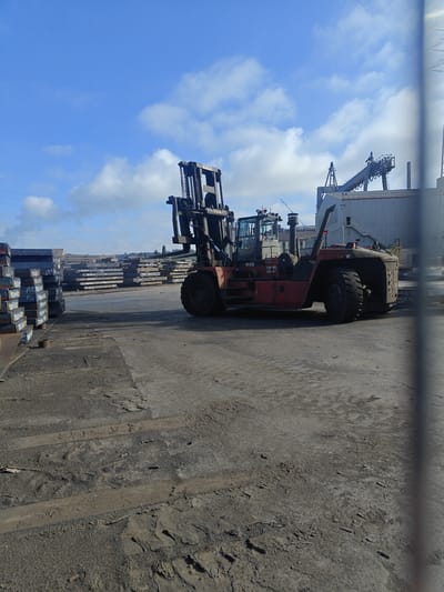 An outdoor scene in the city of Charleroi, Belgium, depicts a large, red forklift truck situated on a dusty, concrete surface. The forklift is positioned in the foreground to the right, with its forks raised and extended. In the background, stacks of metal plates are visible, suggesting a industrial or storage area. Beyond the metal stacks, a large building complex with a loading crane is present. The sky above is blue with scattered white clouds. The overall environment appears to be an open-air industrial yard.