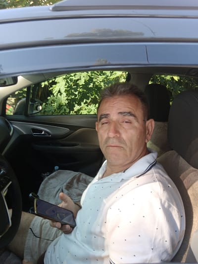 A man sits in the driver's seat of a car, looking towards the camera. He is wearing a white polo shirt with small dark triangular patterns and a blue and white striped collar. He is holding a smartphone in his right hand. The car interior is visible, including a portion of the steering wheel, the dashboard, and the passenger seat. Green foliage is visible through the car window, suggesting an outdoor setting. The lighting indicates it is daytime. No specific details about the "Municipal Unit of Vocha, Greece" are discernible from the image itself.