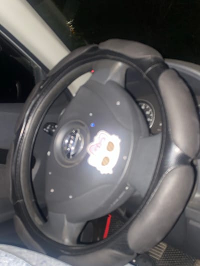 The image shows a close-up view of a car's steering wheel with a protective cover. A sticker with a cartoonish white cat face is affixed to the center of the steering wheel. The background is dark and blurry, suggesting the photo was taken at night or in low-light conditions. Some part of the car's dashboard and interior are visible. The location is specified as Uchaly, Russia, but there are no visual cues in the image to confirm this location.