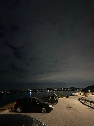 The image captures a night scene along a waterfront in Anatoliki Attiki, Greece. The perspective is from a paved road, looking out over a body of water towards a distant coastline.

In the immediate foreground and to the right, a paved road is visible, where several cars are parked facing away from the viewer. At least three dark-colored cars, possibly hatchbacks, are clearly seen, with their wheels and parts of their bodies illuminated by ambient light. A metal guardrail runs alongside the road, separating it from the water. The road curves gently to the right in the distance, where more streetlights are visible.

The mid-ground is dominated by a calm body of water, likely the sea or a large lake, which reflects numerous bright lights from the opposite shore. These reflections stretch across the water, creating shimmering vertical streaks of light. On the distant landmass across the water, a scattering of artificial lights indicates a populated area.

The sky, occupying the upper left portion of the image, is dark and overcast, with visible cloud formations. The low light conditions suggest it is nighttime. There are no prominent human figures, although vague shapes might be people near the distant lights across the water. The overall atmosphere appears tranquil and serene, characteristic of an evening waterfront view. There is no visible text in the image.