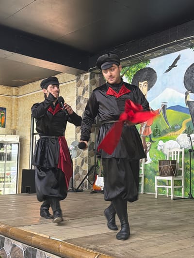 In Kveda Chkhutuneti, Georgia, two individuals are performing on a wooden stage in front of a mural depicting a rural Georgian landscape. The person on the left, wearing a black traditional outfit with red accents and a black cap, is holding a microphone and speaking. The person on the right, similarly dressed in black with red trim and a black cap, is holding a flowing red fabric, possibly for a dance. The mural behind them features stylized figures, sheep, a bird in flight, and a horn. To the left of the stage, a black speaker is visible, and to the right, a white chair with a drum on it and a portion of an electric guitar are present. The lighting suggests an indoor setting, possibly a cultural center or restaurant.