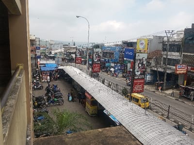 An elevated perspective captures a bustling street scene. A covered walkway dominates the foreground, lined with vendor stalls. Parked motorcycles fill a lot to the left. Across the walkway, a street is lined with buildings displaying various advertisements and storefronts, including "VIVO" and "Alfamat." A yellow vehicle traverses the road. Above the buildings, a cloudy sky completes the urban landscape.