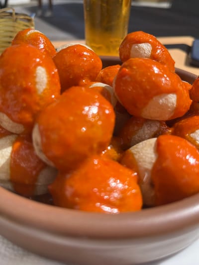 A close-up image of a bowl of papas arrugadas (wrinkled potatoes) with mojo rojo (red sauce) in Agüimes, Spain, at the coordinates 27.907 latitude and -15.447 longitude. The potatoes are piled high in a terracotta bowl and are coated in a thick, bright red sauce. The bowl is partially filled with the potatoes, and the sauce is visible on the rim of the bowl. The image is focused on the potatoes and sauce, with the background blurred out of focus.