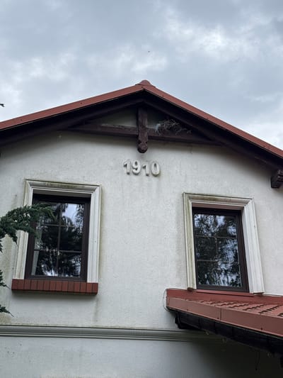 The image shows the exterior of a building with a white stucco facade and a red tile roof. Two windows with dark frames are visible on the upper story. Above the windows, the number "1910" is mounted on the facade. A bird's nest is present on a wooden beam near the peak of the roof. The sky is overcast. The location is Albertowsko, Poland.