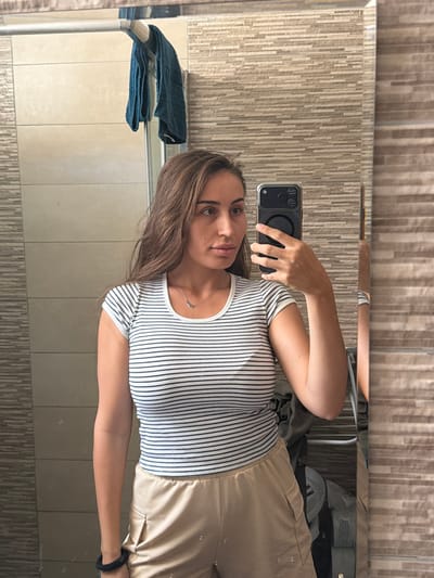 In a bathroom setting in Dromolaxia - Meneou Municipality, Cyprus, a young woman is taking a mirror selfie. She has long brown hair and is wearing a striped t-shirt and light-colored shorts. She is holding a smartphone in her right hand to capture the image. The bathroom features tiled walls and a towel hanging on a rack. The overall scene is casual and domestic.