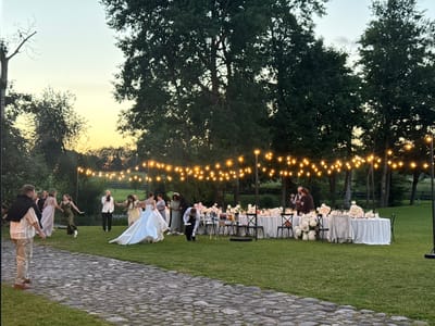 In the city of Ilyanskiy selskiy Sovet, Belarus, an outdoor wedding reception is underway at dusk. The scene is illuminated by a string of warm, glowing lights overhead, casting a festive ambiance. A cobblestone path leads the eye towards a grassy expanse where a wedding party is gathered.
In the foreground, a man in a light-colored shirt and khaki pants stands with his back to the camera, looking towards the reception. Further in the background, a bride in a flowing white gown is captured mid-motion, her dress fanned out around her. Several guests, dressed in various formal attire, are scattered across the lawn, some appearing to dance or mingle.
A long, white-draped table laden with floral arrangements and place settings is set up under the string lights, indicating a formal dining area. Chairs surround the table, some occupied by guests. In the distance, lush green trees and foliage frame the scene, with a hint of the sunset's warm hues in the sky. The overall atmosphere is one of celebration and merriment.