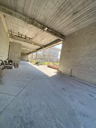 The image depicts an interior view of a construction site or unfinished building. The floor is made of concrete slabs, and the walls are a mix of concrete and light-colored brick. The ceiling is also concrete, with exposed metal beams and pipes. Natural light streams in from an open area at the far end, illuminating the scene and casting long shadows. In the background, industrial structures and greenery are visible outside. There are some construction materials and equipment scattered around, including wooden pallets with cylindrical objects on the left side of the frame, and a rusted cart with metal components in the distance. A person wearing an orange vest is standing near the cart. The overall impression is one of an industrial or commercial space in its raw, early stages of development. The specific location in Police, Poland, is not discernible from the visual information provided.
