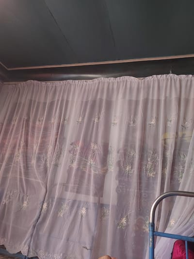 The image depicts an interior scene with a dark teal ceiling composed of wooden planks. A thin wooden trim separates the ceiling from the wall, which is covered by a sheer, light pink curtain. The curtain is adorned with delicate white floral embroidery. The curtain hangs from a rod and is partially drawn, revealing a blurred background that suggests a window or another room. To the right of the curtain, a portion of a blue metal bed frame is visible. The lighting appears to be artificial and ambient, casting soft shadows. There are no people or clear actions taking place in the frame. The scene is domestic and appears to be in a residential setting, possibly a bedroom. The location is identified as Karkidon, Uzbekistan.