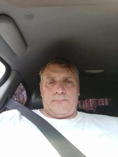 A medium-shot, forward-facing selfie of a middle-aged Caucasian man is captured inside a car. The man is wearing a white t-shirt and a seatbelt is visible across his chest. The interior of the car is dimly lit, with the car's ceiling and part of the dashboard visible. Through the rear window, a forest with tall trees can be seen, suggesting the car is either parked or driving near a wooded area. The man's face is looking directly at the camera with a neutral expression. The image appears to have been taken during the daytime, as indicated by the natural light filtering into the car and illuminating the trees. The location is identified as Klapkalnciems, Latvia.