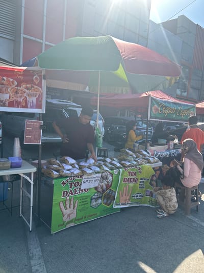 In Makassar, Indonesia, on a sunny day, a street food stall operates under a multi-colored umbrella. The stall sells "Krupuk Toa Daeng" and "Crepes Crepes". A vendor in a maroon shirt stands behind a table laden with bagged snacks labeled "Krupuk Toa Daeng", some of which display the price "Rp. 20.000". A sign to the left advertises a "Snack Tray" and provides a "Daftar Menu". Adjacent to this stall, another stall is visible, with a sign that reads "Crepes Crepes 152". Several people are present: a woman in a yellow shirt serves customers at the crepes stall, a woman in a pink headscarf and shirt sits on a stool looking at her phone, and a child in a patterned outfit crouches on the ground. A man in an orange shirt stands to the right of the crepes stall. The background shows buildings and parked cars. The ground is paved concrete, with sunlight casting shadows.