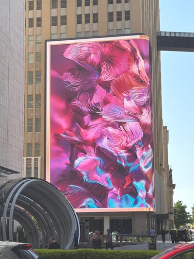 The image shows a large, vibrant digital billboard displaying abstract art with swirling pink and blue patterns. The billboard is mounted on the side of a tall, beige building with numerous windows. To the left of the billboard, a dark, vertical structure resembling a tall antenna or spire rises above the building. The scene appears to be an urban environment, with other buildings and foliage visible in the background. A curved, metallic walkway structure is visible in the lower right corner of the image, with a few people walking on it and nearby. The weather appears to be sunny, suggesting it's daytime. The overall atmosphere is modern and bustling, typical of a city. There is no visible text in the image.