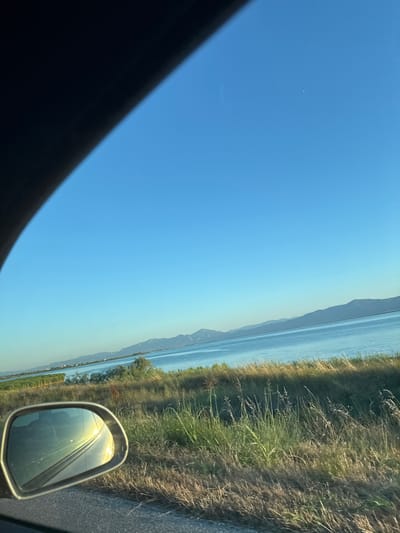 The image captures a scenic view from a car window looking out towards a large body of water and distant mountains under a clear blue sky. The foreground shows a portion of the car's interior, specifically the top of the dashboard and a side-view mirror. The side-view mirror reflects the bright sunlight and the road ahead, creating a lens flare effect.
The environment beyond the car consists of dry, tall grass and vegetation along the roadside, leading to the expansive blue water. The water appears calm with a slight shimmer from the sunlight. In the distance, a range of low, hazy mountains lines the horizon. The sky is a vivid, cloudless blue, suggesting a clear and sunny day.
There are no people visible in the image, and no specific city or location in Greece is identifiable from the visual information provided. The scene suggests a drive along a coastal or lakeside road.