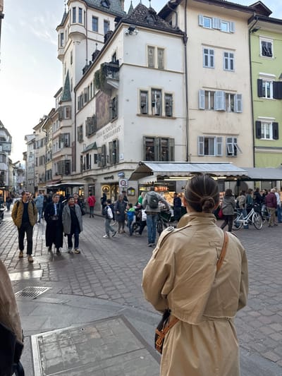 The image depicts a bustling cobblestone street in Bolzano, Italy, characterized by historical architecture under soft, possibly late afternoon or overcast, lighting.

In the immediate foreground, facing away from the camera, is an individual wearing a beige, double-breasted trench coat with a cropped design at the waist and a brown leather shoulder bag slung across their back. Their dark hair is pulled back into a neat bun.

The mid-ground features a lively pedestrian area with numerous people. To the left, a group of three individuals walks towards the viewer; the closest wears a yellow puffer jacket, dark trousers, and dark shoes. Further into the mid-ground, a market-like setup with white awnings and visible food displays occupies the right side of the street. Several individuals are gathered around this area. A man in a grey hoodie and jeans stands next to a bicycle in front of the market stall. Two white bicycles are parked nearby.

The background is dominated by multi-story European buildings. A prominent white corner building, identified by a street sign as "CASA AL TORCHIO JORGGLHAUS," features a tall, slender octagonal tower with a dark conical roof, bay windows, and traditional green shutters on multiple floors. Some windows have decorative wrought iron elements, and a balcony on the second floor is adorned with green plants. Adjacent to this, a light yellow building and a pale green building extend to the right, both featuring multiple windows with light-colored shutters, many of which are open. To the left of the corner building, the street recedes into a narrower passage lined with similar historic buildings, one of which displays a clock. The sky above is a pale blue with minimal cloud cover.