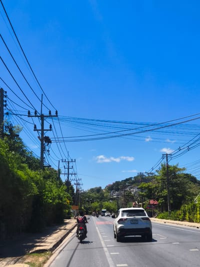 This is a vertical, eye-level shot of a road in Baan Thong Po, Thailand, on a bright, clear day. The sky is a vibrant blue with a few scattered white clouds. Utility poles with a dense network of black wires run along the left side of the road, interspersed with lush green trees and foliage. A sidewalk is visible on the left, partially shaded by the trees.
The road itself is paved and marked with white lines. Several vehicles are present: a white SUV is in the foreground on the right side of the road, and a person on a black motorcycle is in the left lane ahead of the SUV. Further down the road, other vehicles and people are visible, suggesting a moderate level of traffic. In the background, the road curves uphill towards a hillside covered in buildings and more greenery, characteristic of a tropical urban environment. The overall impression is one of a sunny day in a busy, tropical town.