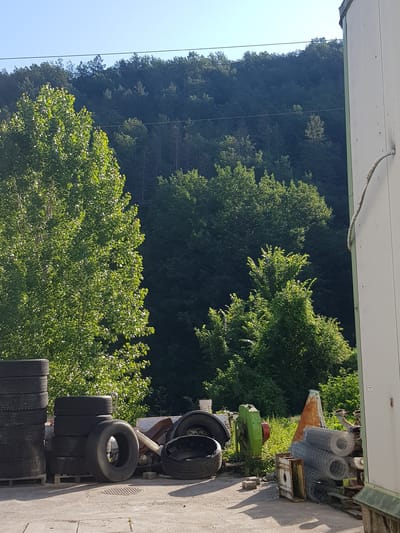 This is an exterior, eye-level, medium shot of a storage area located in Lomets, Bulgaria. The scene is dominated by a densely wooded hillside in the background, with a bright blue sky above. In the foreground, to the left, is a tall stack of black tires. To the right of the tires, there are more tires, some stacked and one lying flat with its inner cavity visible. Behind the tires, to the right, is a tall, white rectangular structure, possibly a shipping container or building. A thick, beige rope is attached to the side of this structure and trails upwards. Further to the right and slightly behind the white structure, there are rolls of wire mesh stacked. In the middle ground, various pieces of machinery and discarded items are scattered, including a green machine with red circular components and some orange cones. The ground is a light-colored paved surface, with shadows cast by the objects. Lush green trees and foliage are visible throughout the scene, particularly in the middle and background. There are no people visible in the image.
