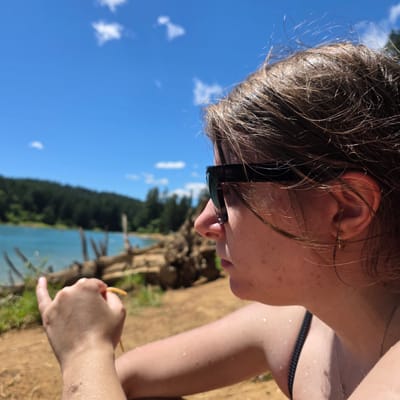 A medium shot shows a person wearing black sunglasses and a black bikini top, looking out at a blue lake bordered by a dark green forest under a bright blue sky with scattered white clouds. The person's right hand is in the foreground, holding a yellow object. The person's skin is wet, with small water droplets visible on their arm and neck. The location is Cherry Grove, United States.