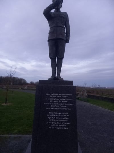 A statue of a soldier saluting stands beside a black monument engraved with a poem. The poem is printed in both French and English. The French text begins, "Ils ne vieilliront pas comme nous," while the English version starts, "They shall grow not old." The poem discusses themes of remembrance and enduring respect for those who have passed. The setting includes a grassy area, trees in the background, and a cloudy sky. There is a bench visible in the distance.