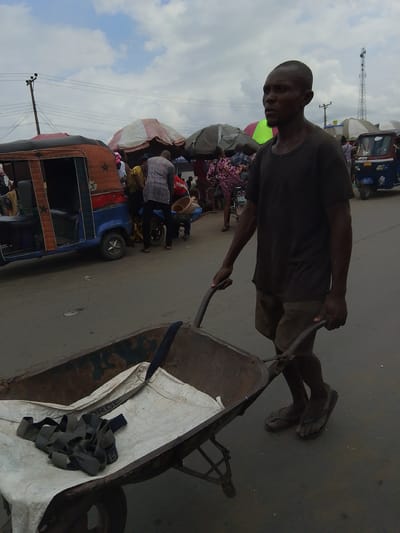 A man is pushing a wheelbarrow in a busy outdoor market. He is wearing a dark t-shirt and shorts, and flip-flops. The wheelbarrow is filled with a white tarp and some straps. In the background, there are several other people, some standing and some walking, and a few small vehicles that appear to be auto-rickshaws, suggesting this is a market area. There are also market stalls with umbrellas. The sky is cloudy, indicating it might be daytime, possibly mid-morning or afternoon. The overall scene depicts a bustling commercial environment. The text "FORGE" is visible on one of the straps in the wheelbarrow. The ground appears to be paved, possibly a road or a section of the market. The man's posture suggests he is actively engaged in his task. The location context is Yenegoa, Nigeria.