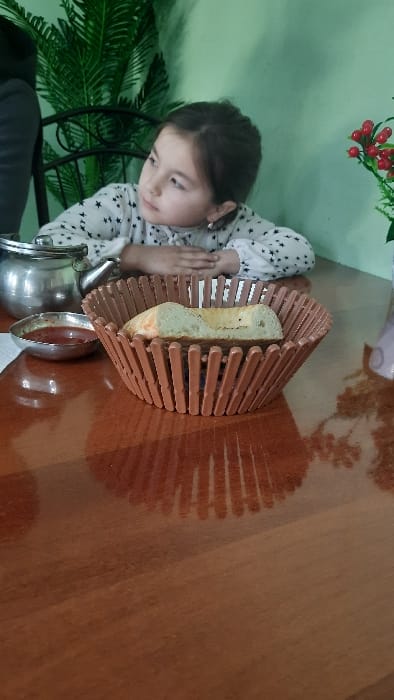 The image displays a young girl seated at a glossy, reddish-brown wooden table within an establishment in Yangiariq, Uzbekistan. The girl, estimated to be between 5-8 years old, has dark hair pulled back and wears a white long-sleeved shirt patterned with black stars and a light-colored bow at the neckline, along with small silver-colored stud earrings. Her hands are clasped together on the table, and her attention is directed towards the upper left, off-camera.
On the table, from left to right, are a small, silver-toned metallic teapot, a small silver-colored bowl containing a red liquid substance (likely a sauce or dip) resting on a partially visible white napkin, and a brown, slatted basket holding a slice of white bread. A dark-colored sleeve, belonging to another individual, is partially visible on the far left. The background features a light mint green wall, an artificial dark green plant with broad leaves behind the girl to her left, and another artificial plant with red berries on green stems to her right. Reflections of the basket and other elements are discernible on the table's polished surface.