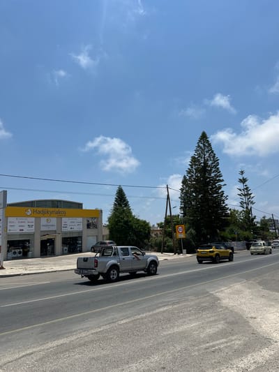 This media file captures a street scene in Mesogi, Cyprus, on a bright, sunny day. The perspective is from the side of the road, looking towards buildings and a tree-lined area. Dominating the left side of the frame is a tall, triangular pine tree, its branches reaching towards a clear blue sky dotted with fluffy white clouds.
In the foreground, a silver pickup truck with its doors open is parked on the side of the road. A person's arm is visible reaching out of the driver's side window. Further down the road, a bright yellow car is also parked. In the background, several buildings are visible, with signage indicating shops. One prominent sign displays "Hadjikyriakos" in large yellow letters, along with brand names like "BOSCH," "SHARP," "AEG," and "LG," suggesting an appliance store.
A speed limit sign indicating "50" with "Pafos" written below is also visible on the side of the road. The overall scene depicts a typical day in a Cypriot town, with cars parked along the street, shops open for business, and lush greenery providing a contrast to the urban elements. The bright sunlight and clear sky suggest it is midday.