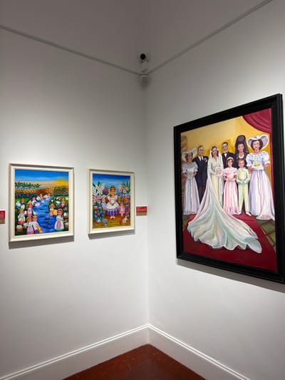 The image depicts an art exhibition room, likely a gallery, with white walls and a dark wooden floor. Three paintings are displayed on the walls. On the left, there are two framed paintings with white borders. The first painting, to the left, shows a scene with people in a natural setting, possibly near a body of water, under a bright sky. The second painting, to the right, features a figure in the center surrounded by flowers. To the right of these, and larger in size, is a framed painting with a black border. This painting portrays a formal gathering, appearing to be a wedding party, with several individuals dressed in elegant attire. A bride in a white gown with a long train is the central focus. The paintings are hung at eye level, and a security camera is visible on the ceiling in the upper left quadrant of the image. Red labels with text are present next to the two smaller paintings, and a similar label is to the left of the large painting. The lighting is bright, illuminating the artwork and the room. The environment suggests a public space dedicated to showcasing art.