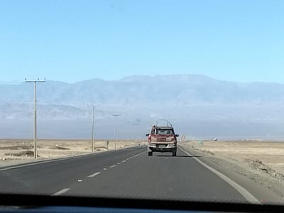 The media file captures a view from a vehicle traveling on a paved road through a desolate, arid landscape. In the foreground, the road stretches out, marked by white dashed lines and solid white lines at the edge. The landscape to the sides of the road is dry and barren, with sparse, low-lying vegetation and rocky terrain. Several utility poles are visible along the roadside, supporting power lines against a clear, pale blue sky. In the middle ground, a red SUV is driving away from the viewer on the same road. The SUV appears to be carrying items on its roof. In the distance, a range of hazy, bluish mountains forms the horizon. The overall scene suggests a sunny day with excellent visibility, typical of a dry, desert environment. The location is identified as Pozo Almonte, Chile, which is known for its desert climate. There is no visible text in the image.