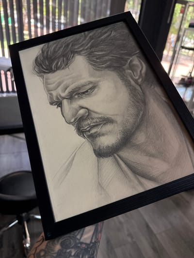 This is a close-up, angled shot of a framed pencil drawing of a person's face, possibly a portrait, resting on a surface. The drawing is in black and white, with detailed shading to depict features like hair, an eye, nose, and mouth. The subject appears to be sleeping or in a state of repose, with closed eyes and a relaxed expression. The drawing is enclosed in a thin, black frame.
In the background, there are indications of an indoor setting, with horizontal blinds creating patterns of light and shadow. Through the blinds, glimpses of greenery and the bright light of daytime are visible, suggesting the drawing is positioned near a window. The floor appears to be wood-paneled. To the right of the frame, a tattooed arm is partially visible, with intricate designs. The overall impression is a personal display of artwork within a domestic or studio environment. The context provided by "Campillo del Moro, Spain" places this scene in a specific geographical location. No visible text is present in the image.