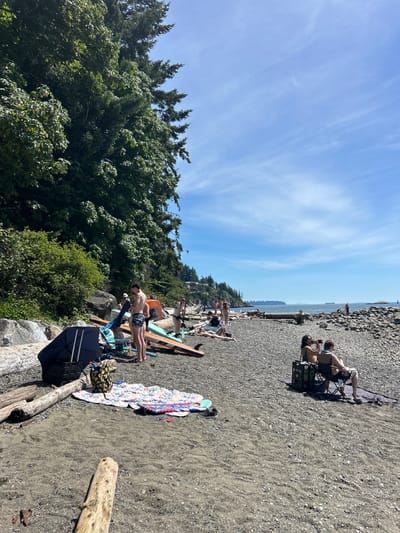 This image depicts a sunny day at a pebble beach, likely located in West Vancouver, Canada, given the dense evergreen trees and the coastal landscape. Several people are present, enjoying various beach activities. In the foreground, on the sand, lies a colorful patterned blanket, a closed dark blue umbrella, and some driftwood logs. Further back, on the pebble shore, a group of individuals are gathered near several paddleboards. One man is standing next to the paddleboards, while others are seated or lying down on the beach. Two people are seated in chairs in the middle ground, one of whom is holding a camera and looking out towards the water. The ocean stretches out to the horizon under a bright blue sky with scattered white clouds. The shoreline is rocky, with more driftwood scattered along the beach. In the distance, several ships are visible on the water, and the coastline is lined with trees and houses.