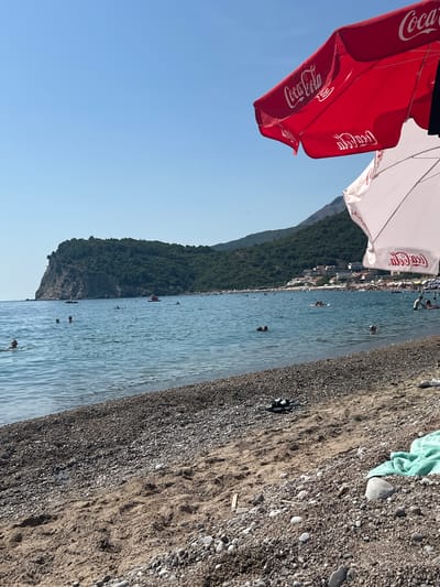 This is an eye-level, outdoor, medium shot of a pebbly beach leading to the Adriatic Sea. The water is a clear blue, with small waves and several people swimming and wading. A large, forested island with a rocky cliff face is visible in the distance, along with buildings and a densely packed beach with numerous sunbathers. Two large Coca-Cola branded beach umbrellas, one red and one white, are in the foreground, casting shadows on the sand. On the beach, a light blue towel and a pair of dark sandals are visible. The sky is clear and blue. This scene is located in the city of Đurovići, Montenegro.