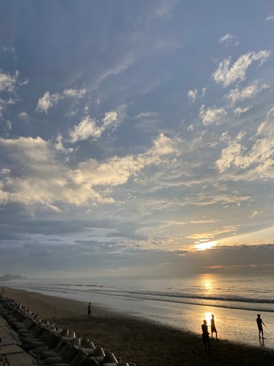 The image depicts a serene beach scene at Bateetimoh, Indonesia, during sunrise. The sky is filled with a blend of light and dark clouds, with the sun casting a warm, golden glow as it peeks through the clouds near the horizon. The ocean is calm with gentle waves lapping at the shore. On the wet sand of the beach, three figures are visible, silhouetted against the bright sun, appearing to be casually interacting. In the foreground, a concrete barrier wall runs along the edge of the beach, with a person standing near it further down the shoreline. The overall atmosphere is tranquil and peaceful.