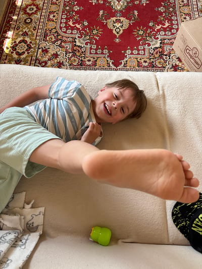 The image shows a young boy lying on a couch in what appears to be a residential setting. The boy is wearing a striped t-shirt and light-colored shorts. His left leg is extended towards the camera, with his foot in sharp focus. He is smiling broadly and looking up. The couch is covered with a beige fabric. Behind the couch, a red oriental rug with a floral pattern is visible on the floor. A cardboard box with Cyrillic lettering is also present on the rug. On the couch, near the boy's foot, is a small green toy. In the lower left of the image, a white blanket with a pattern of cartoon animals is visible. In the lower right, a black and yellow patterned object, possibly a hat or bag, is partially visible. The scene is illuminated by natural light, likely from a window. There is no information to indicate the location in the city of Радваніцкі сельскі Савет, Belarus.