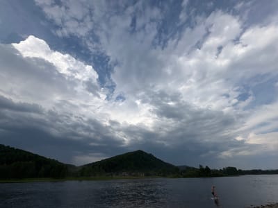 A scenic view of a wide, calm river is captured under a dramatic, cloud-filled sky. The sky is a mix of dark, brooding storm clouds and brighter, white cumulus clouds, suggesting a day of changing weather, possibly near dusk. The river is dark and reflects the sky's moody tones. On the far bank of the river, a dense, green forest covers rolling hills and a prominent, conical mountain. A few scattered buildings are visible amongst the trees at the base of the mountain.
In the foreground, a lone person, wearing dark shorts, is wading into the river from a rocky shore. Their posture suggests they are either entering or exiting the water, perhaps for a swim or to cool off. Their pale skin stands out against the dark water and the muted landscape. Further out in the river, two other figures are partially visible, also in the water, suggesting this is a popular spot for recreation. The overall mood is one of serene, wild beauty, tinged with the dramatic atmosphere of an approaching or passing storm. The location is identified as Mezhdurechensk, Russia, known for its natural landscapes and rivers.