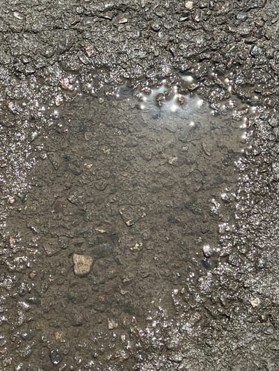 The image shows a close-up view of a puddle on asphalt in Qoyankus, Kazakhstan. The asphalt is rough and composed of small dark gray stones. The puddle is in the center of the frame, reflecting sunlight. The edges of the puddle are irregular and are surrounded by wet asphalt that sparkles with light reflections. Several small stones are visible both in the dry asphalt and submerged in the water. There are no people or animals visible in the image. The scene appears to be an outdoor environment, likely a street or parking lot, after rain.