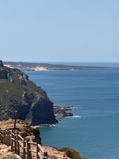 A vertical, eye-level, outdoor, wide shot captures a sunlit, coastal landscape in Colares, Portugal. The scene is dominated by the vast expanse of the deep blue ocean, which occupies the right two-thirds of the frame. The water is calm with subtle ripples and a few whitecaps suggesting a gentle breeze.

In the foreground, a rustic wooden fence made of vertical posts and horizontal rails runs along the edge of a rocky, reddish-brown cliff. The cliff face is uneven and sparsely covered with low-lying green vegetation. A tall, weathered wooden post stands near the fence, leaning slightly to the left.

To the left of the frame, a steep, rocky cliff descends dramatically towards the ocean. The cliff is layered with dark, angular rock formations and patches of dry, light-brown grass and hardy green succulents. The vegetation on this cliff is sparser than on the foreground cliff.

In the middle ground, the ocean extends to a distant, hazy coastline characterized by sand dunes and low-lying green scrub. Further in the distance, a faint, dark blue horizon line separates the sea from a pale blue sky. The overall atmosphere is one of natural beauty and a sense of remote, rugged coastline.