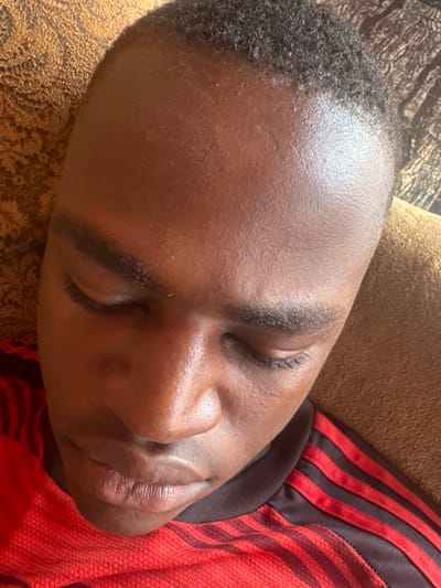 A close-up, high-angle image depicts a person of African descent, with their eyes closed, appearing to be asleep or at rest. The individual has short, dark, curly hair, dark skin, prominent eyebrows, a broad nose, and closed lips. They are wearing a red shirt with horizontal black stripes across the shoulders and upper chest. The background on the left features a patterned light brown or gold fabric, while the right side shows a plain tan or light brown upholstered surface. The scene is located in Umuoye, Nigeria.