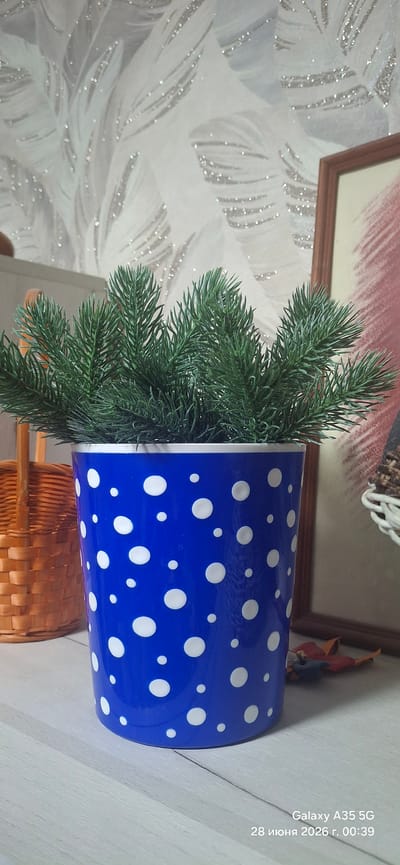 A close-up, vertically oriented shot captures a small, artificial evergreen tree with vibrant green foliage. The tree is placed inside a bright blue pot adorned with a white polka dot pattern. The pot, in turn, rests on a woven, light brown basket. In the background, a wall with a light-colored, textured wallpaper featuring feather-like designs and subtle glitter accents is visible. A wooden picture frame is also present, hanging on the wall behind the pot. The overall scene suggests an indoor setting, possibly a home or decorative display. A timestamp in the upper right corner reads "Galaxy A35 5G 28 июня 2026 г. 00:39", indicating the image was taken in Russia, specifically in Belorechensky, at a late hour on June 28, 2026. There is no visible weather information or emotional cues conveyed by the image.