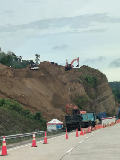 This image depicts a construction site located on a steep, muddy embankment in Sihomlhok, Indonesia. Several heavy machinery vehicles, including excavators and dump trucks, are actively engaged in earthmoving operations. One excavator is positioned at the top of the embankment, actively digging, while another is on the slope. Two dump trucks are parked at the base of the embankment. A small tent with signage reading "Safety is our spirit" is set up near the construction equipment. Traffic cones and a guardrail are present along the road in the foreground, indicating a work zone or hazard area. The sky is overcast, suggesting potentially damp or rainy weather.