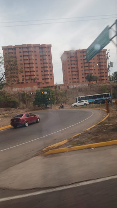 The image shows a street scene in Puerto La Cruz, Venezuela, featuring two tall, orange-colored, multi-story residential buildings. A road curves in front of the buildings. A red car is on the road, and a white bus is parked near the edge of the road. A pedestrian or cyclist is also visible near the bus. A street sign hangs over the road in the distance. The road has yellow painted edges and a white dividing line. The sky is overcast.