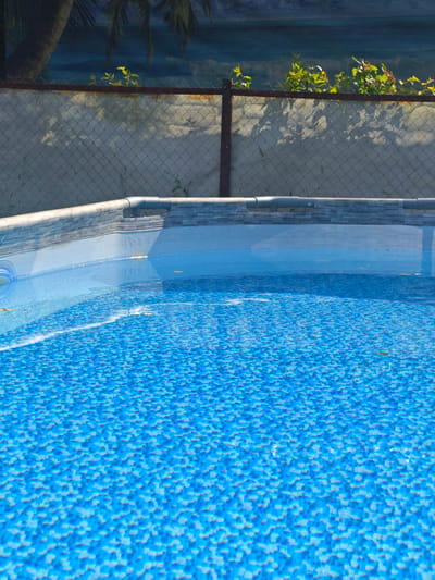 This media file depicts an above-ground swimming pool in a backyard setting. The pool has a blue, tiled pattern at the bottom and is filled with clear water. The edges of the pool are lined with a faux stone material. In the background, a chain-link fence separates the pool area from a more vegetated area. Beyond the fence, palm fronds are visible, suggesting a tropical or subtropical climate, although the location context indicates Хлевино, Russia. Shadows cast by foliage are visible on the fence and the wall behind it, indicating it is daytime. There are no people or active events depicted. The overall mood is tranquil and still.
