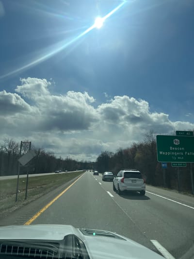 The image shows a highway scene near Exit 41 in the Town of Fishkill, United States of America. The road sign indicates exits for Beacon and Wappingers Falls, which are 1/2 mile ahead. The road appears to be a highway, with multiple lanes and a grassy median. Several cars are visible on the road, including a white SUV and the edge of the vehicle from which the photo was taken. The sky is mostly blue with large, puffy white clouds. The sun is shining brightly, creating a lens flare. There are bare trees bordering the highway.