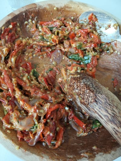In a wooden bowl, a vibrant red chili paste, or sambal, is being prepared. The paste contains visible chili seeds, chopped red chilies, translucent onion slices, and green herbs. A dark wooden pestle rests within the bowl, indicating the ongoing grinding process. A silver spoon also sits in the mixture. The bowl's natural wood grain adds texture to the rustic composition. The overall impression is one of fresh, homemade spice.