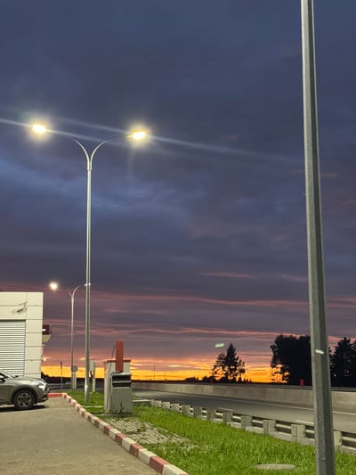This media file depicts an outdoor scene during twilight, likely in Можга, Russia. The sky is filled with dramatic clouds tinged with orange and purple hues, suggesting a sunset or sunrise. Tall streetlights with illuminated bulbs are prominent in the foreground, casting a glow against the darkening sky. A road or pathway is visible, bordered by a curb with red and white markings. To the right, a patch of green grass and a concrete barrier with regularly spaced indentations are present, possibly indicating a roadside or park area. In the background, silhouetted trees are visible against the vibrant sky, and the lights of distant structures can be seen. A portion of a building with a ribbed facade and a security camera is in the lower right corner, along with the rear of a vehicle. The overall atmosphere is calm and serene, with the vibrant colors of the sky being a focal point. The time of day is clearly twilight, given the illumination of the streetlights and the colors in the sky. There is no visible text in the media file.