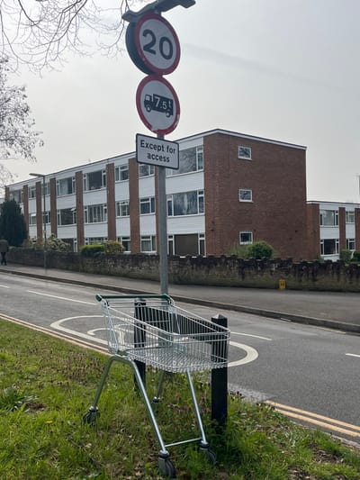 The image displays a street scene within Elmbridge, United Kingdom, under an overcast sky. In the immediate foreground, a silver-colored metal shopping cart with green plastic handles is positioned on a grassy verge, leaning against a black square-profile bollard. The verge exhibits green grass, some dandelions, and small purple-flowered weeds.
To the left, a grey asphalt road features white and yellow line markings, including a semi-circular white marking on the pavement. Further left, a person dressed in dark clothing is seen walking away from the viewer on a sidewalk adjacent to the road, partially obscured by a bare-branched tree.
Centrally located, a grey metal pole supports a street light fixture at its apex and three stacked road signs. The topmost sign is a circular red-bordered white sign displaying "20" in black, indicating a 20 mph speed limit. Directly below it is another circular red-bordered white sign featuring a black truck icon with "7.5t" underneath, denoting a 7.5-tonne weight restriction for vehicles. The bottom sign is a rectangular white sign with a black border, bearing the black text "Except for access."
In the background, a multi-story residential building, likely comprising apartments or flats, presents a facade of alternating brown brick sections and white paneled sections, each containing multiple windows. A low, weathered stone wall with an irregular top edge runs along the front of the property, separating it from the street. Various shrubs and some foliage are visible behind the wall. A small yellow 'H' marker is painted on the pavement near the wall, indicating a fire hydrant location.