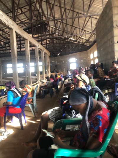 In Nteje, Nigeria, a large indoor space with a high, exposed roof structure of steel beams and support columns houses numerous people seated in plastic chairs arranged in rows. Sunlight streams in through openings in the roof, illuminating the individuals as they are bent over, likely writing or reading, presumably taking a test or examination. The floor appears to be bare and unmarked. The scene suggests a temporary testing center.