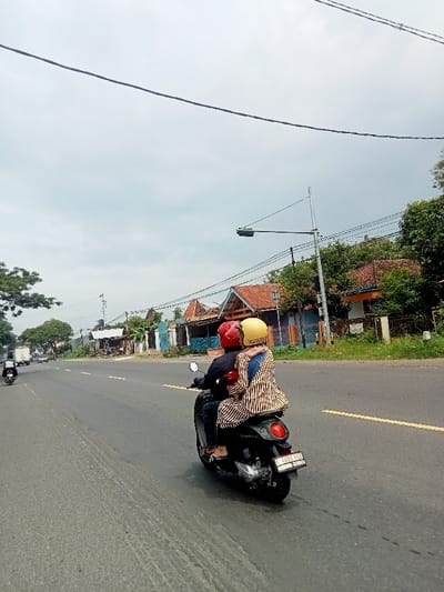 The image shows a man and a woman riding a motorcycle on a paved road in Kecamatan Margorejo, Indonesia. The man is wearing a red helmet and a blue shirt, and the woman is wearing a yellow helmet and a striped shirt. They are riding on a green scooter, and the woman is holding onto the man. There are some houses on the right side of the road, and a couple of cars in the distance. The weather is cloudy, and the road is straight with a faint yellow stripe down the middle. The location is near the coordinates -6.787 latitude and 110.981 longitude, in a residential area. The motorcycle has a license plate that reads "4265 PU". The motorcycle is facing forward, and the man and woman are looking straight ahead.