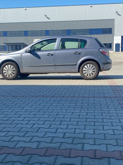 The image shows a side view of a grey hatchback car parked on a paved area. The car is oriented vertically in the frame, with the front facing upwards and to the right. It has tinted windows, silver door handles, and a roof rack. The wheels have silver hubcaps. The car is parked next to a building with a modern facade, characterized by horizontal white metal siding and blue-tinted windows arranged in horizontal bands. The ground is covered with interlocking pavers in shades of grey and a few red ones, forming a patterned walkway or parking area. Shadows cast by the car and the building suggest it is daytime, likely mid-morning or afternoon, with clear weather. The location is identified as Bielany Wrocławskie, Poland. There are no people visible in the image.