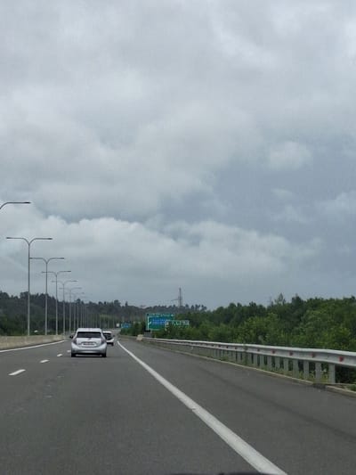 The image depicts a highway scene on an overcast day in Shekvetili, Georgia. A silver hatchback is driving on the right lane of the highway, followed by another car. The highway is flanked by rows of streetlights on the left and a metal guardrail on the right. Lush green trees and vegetation border the highway, with a distant view of what appears to be a power line tower. A green road sign is visible in the distance on the right side of the road. The sky is filled with thick, gray clouds, indicating potentially rainy weather. The perspective is from inside a vehicle looking forward along the road.