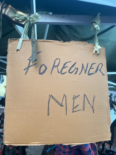 The image shows a roughly cut piece of cardboard with the words "FOREIGNER MEN" written in large, black marker. The sign is hanging from a white metal structure, possibly part of a temporary shelter, by two pieces of tied fabric. The environment around the sign appears cluttered, with possible glimpses of other people and objects blurred in the background. The precise location within Mira-Bhayander, India cannot be determined from the given image.